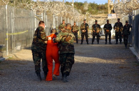 U.S. soldiers force Guantanamo prisoner in orange jumpsuit to walk.
