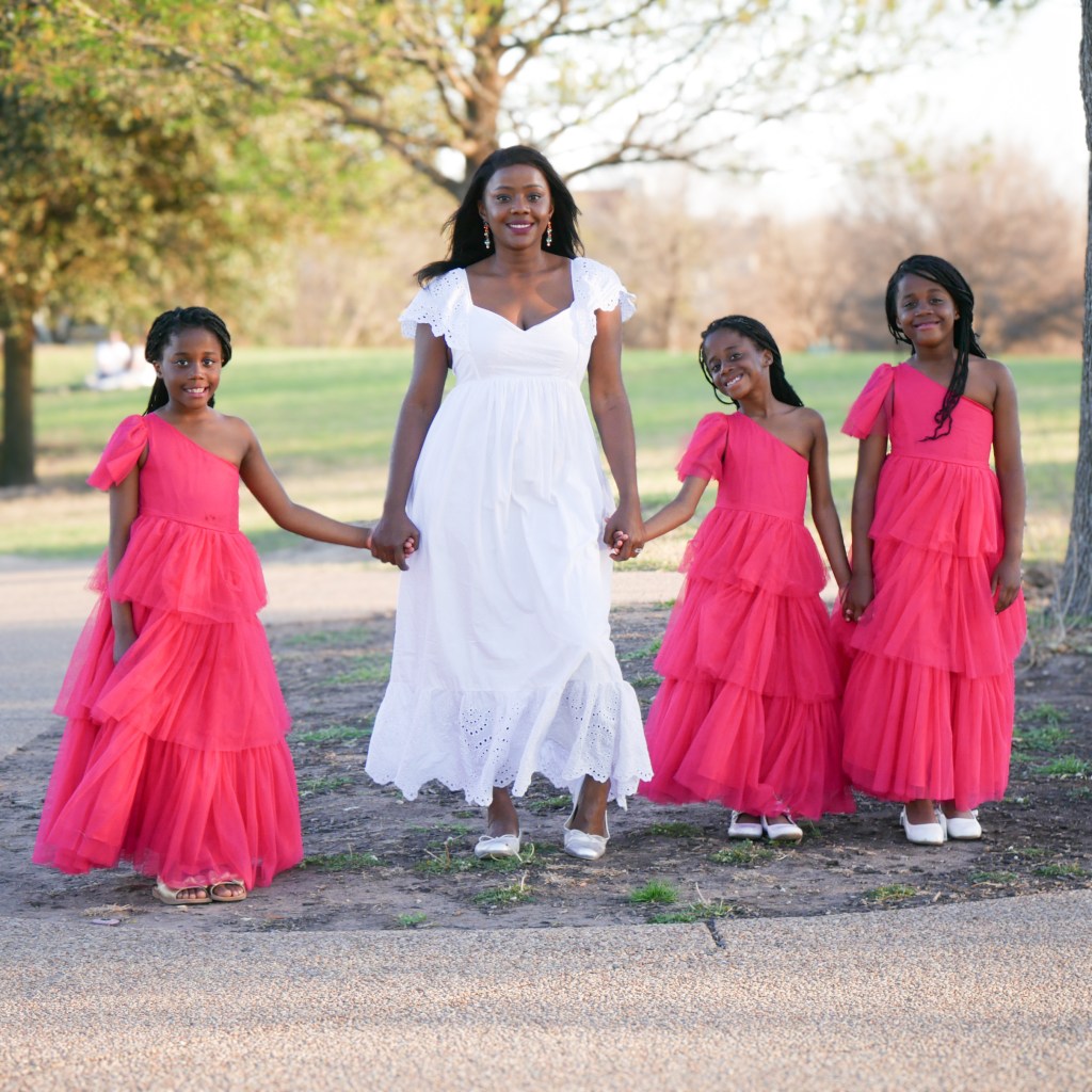 A woman in a white dress stands smiling, holding hands with three young girls in vibrant pink dresses in a park setting, with trees and a grassy area in the background.